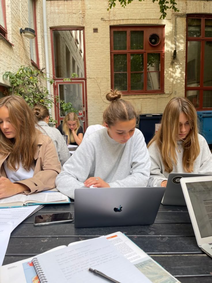 Eine Gruppe von vier Studentinnen lernt gemeinsam an einem Holztisch im Innenhof einer Universität. Sie arbeiten konzentriert an Laptops und mit handschriftlichen Notizen in ihren Büchern – eine produktive Lernsession unter freiem Himmel.