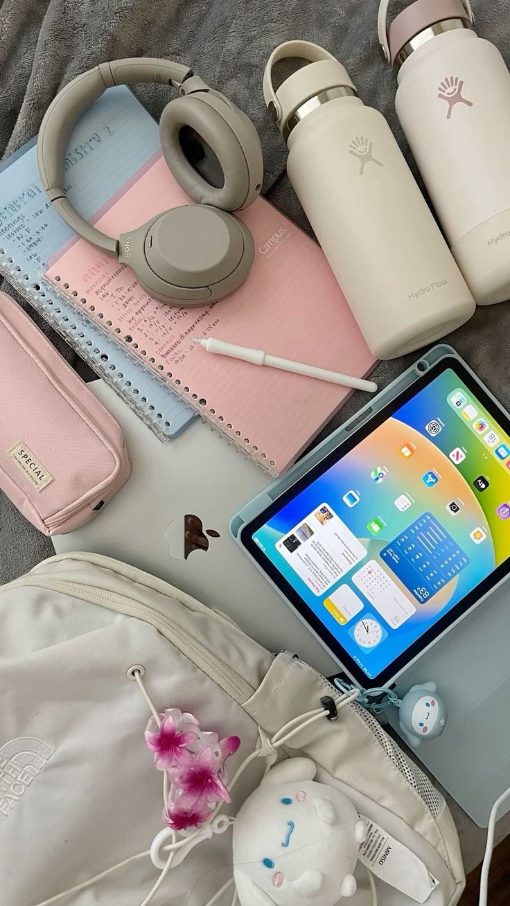 Essentials für den Semesterstart: Noise-Cancelling-Kopfhörer, Notizbücher, iPad, Apple Pencil, Wasserflaschen, Laptop, Federmäppchen und Rucksack auf einer Decke arrangiert.